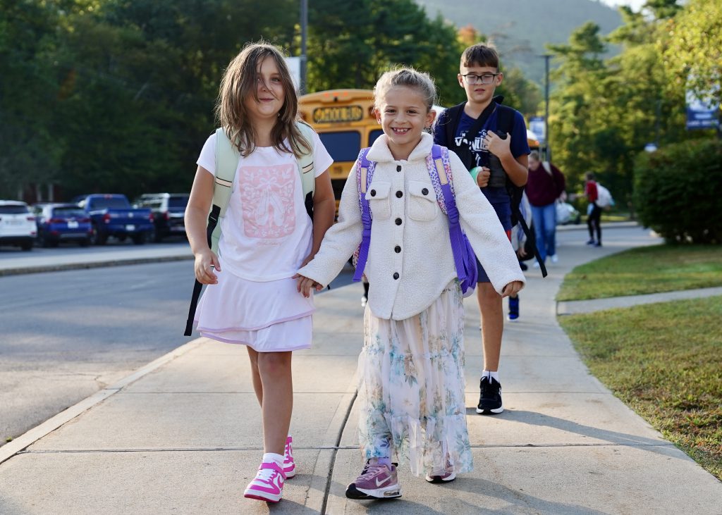 Two girls holding hands and walking with a school bus in the background