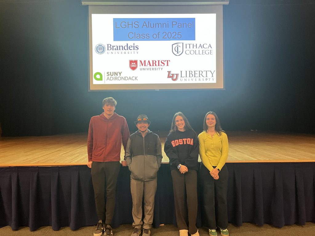 Four former students standing in front of a stage with a sign of college names behind them