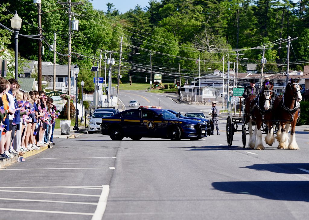 Horse drawn carriage in front of a police car with students holding flags on the side