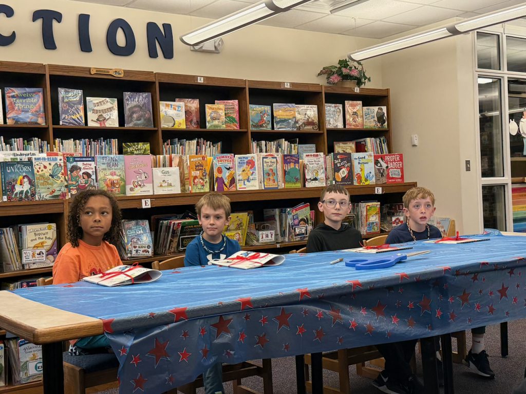 Four students sitting at a table with a tablecloth with books behind them
