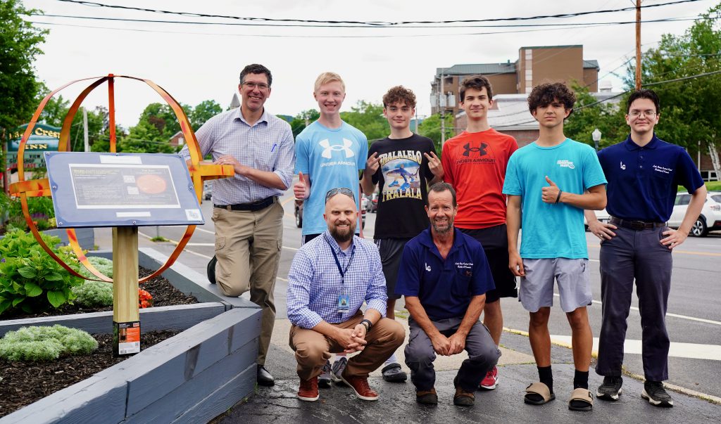 four students and staff standing in front of a welded metal sun that is yellow, orange and red