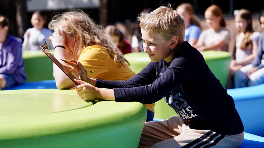 elementary school student in blue shirt holding a clip board outside sitting at a green table